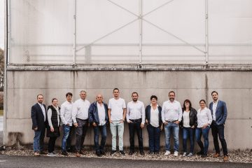 Gruppenszene von 12 Personen in weißen und blauen Shirts, stehend vor einer grauen Wand mit industriellem Hintergrund.