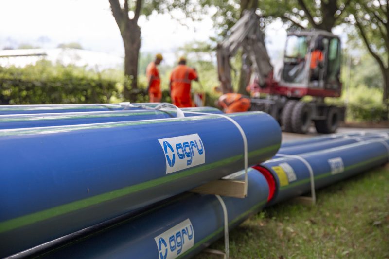 Blaue Rohre liegen im Vordergrund, während Arbeitende in orangefarbenen Overalls im Hintergrund mit einem Gerät beschäftigt sind.