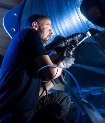 Ein Mechaniker arbeitet konzentriert mit einem Werkzeug an einer großen Maschine, umgeben von bläulichem Licht.
