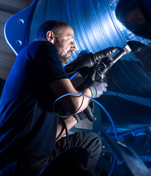Ein Mechaniker arbeitet konzentriert mit einem Werkzeug an einer großen Maschine, umgeben von bläulichem Licht.