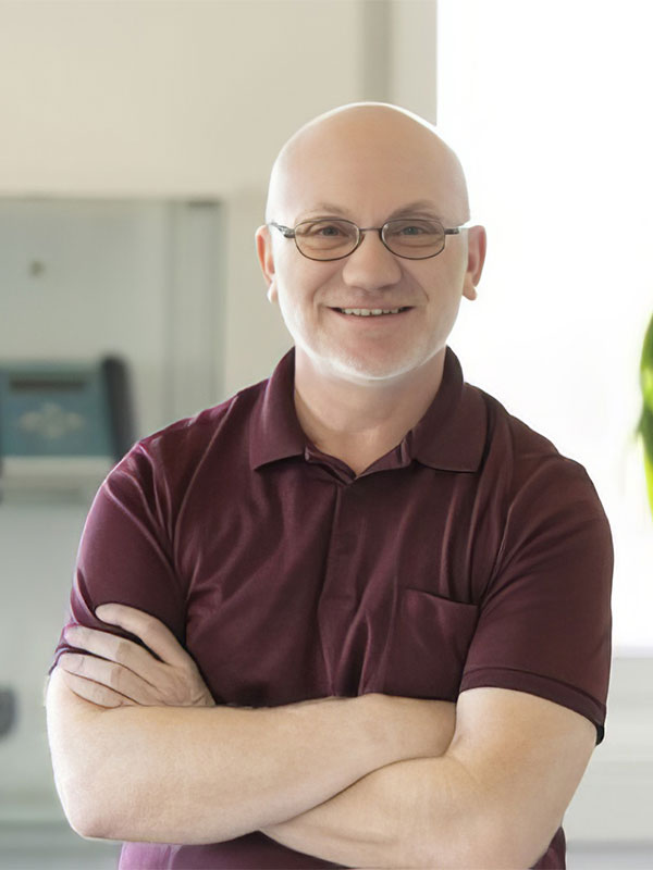 Ein lächelnder Mann mit Glatze und Brille, in einem bordeauxfarbenen Poloshirt, steht mit verschränkten Armen in einem hellen Raum.