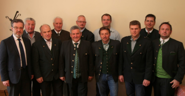 Zehn Männer in Anzügen und Trachtenjacken stehen nebeneinander, lächeln in die Kamera, Gruppenfoto in einem hellen Raum.