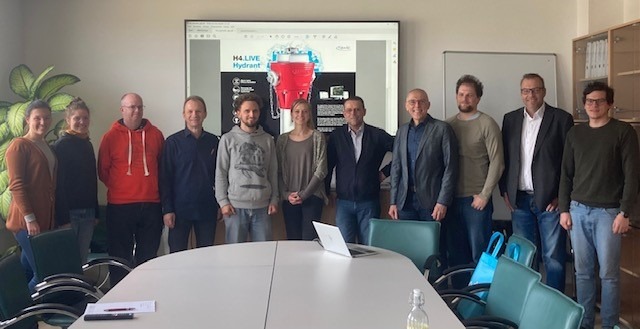 Eine Gruppe von zehn Personen steht in einem Besprechungsraum vor einem Bildschirm mit einer Präsentation.