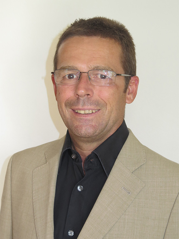 Ein Mann mit Brille trägt einen hellen Anzug über einem dunklen Hemd und lächelt vor einem neutralen Hintergrund.