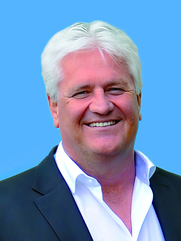 Ein lächelnder Mann mit kurzen, grauen Haaren trägt einen dunklen Anzug und ein weißes Hemd, vor blauem Hintergrund.