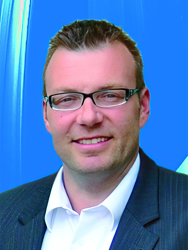 Porträt eines Mannes mit Brille, in Anzug und Hemd, vor einem blauen Hintergrund, lächelnd und selbstbewusst.