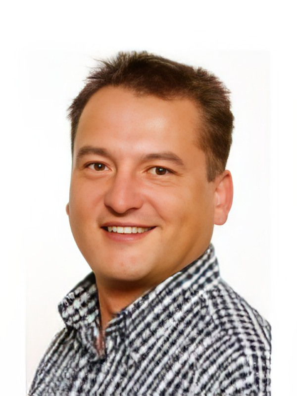 Portrait eines lächelnden Mannes mit kurzem Haar, der ein kariertes Hemd trägt. Heller Hintergrund.